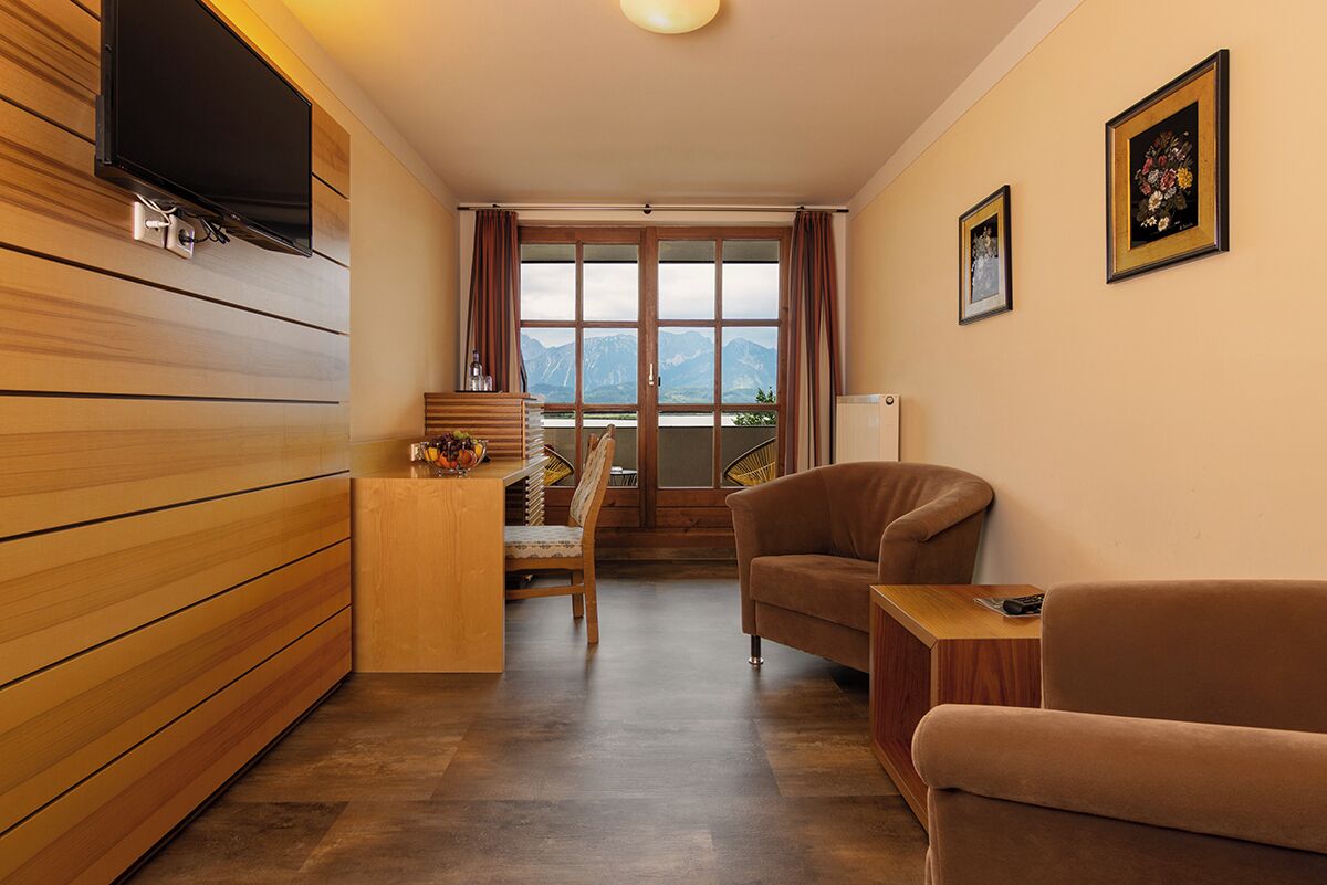 Ein modernes Wohnzimmer mit Holzboden, einer Couch, einem Sessel, einem an der Wand montierten Fernseher und Bergblick durch große Fenster.