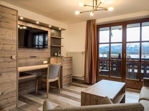 Una sala de estar moderna con un escritorio de madera, una silla cómoda, un sofá y vistas a montañas a través de amplios ventanales.