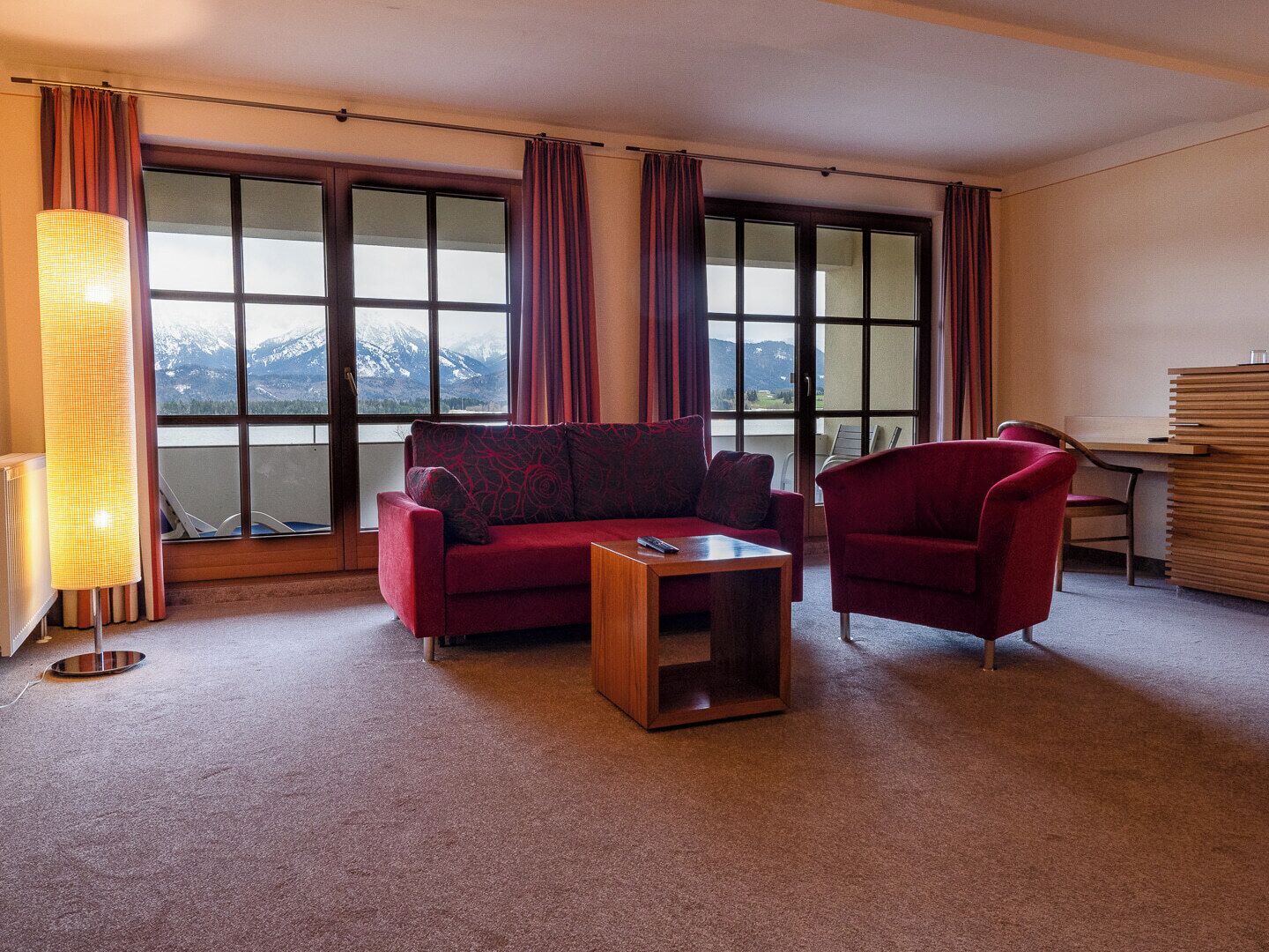 Une pièce avec un canapé rouge, une table basse en bois et un fauteuil rouge. Une grande fenêtre avec des rideaux est visible, et une lampe se trouve sur la gauche.