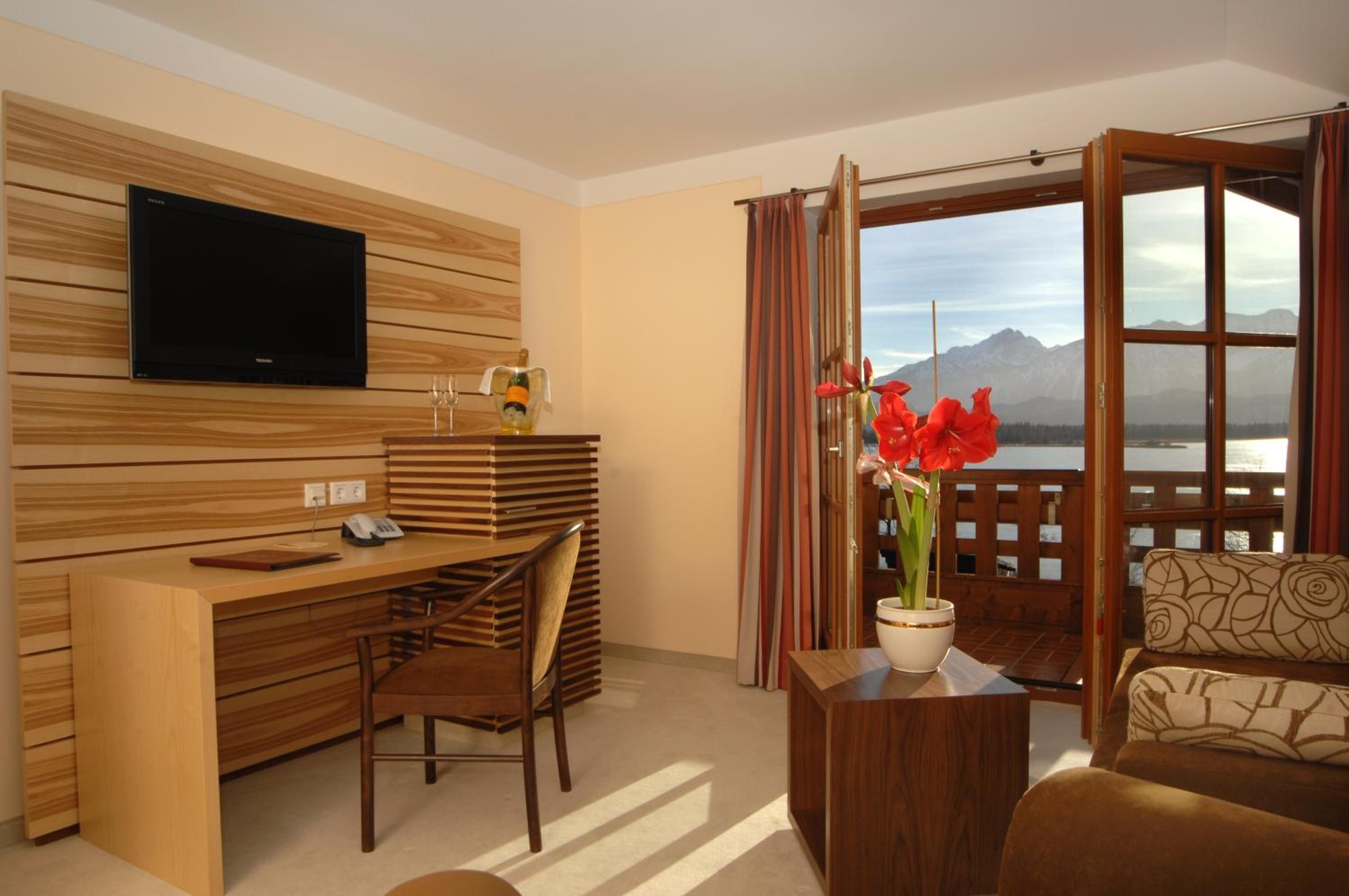Habitación de hotel moderna con pared de madera, televisor de pantalla plana, escritorio con silla, sofá y vistas a montañas y un lago.