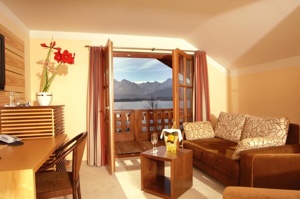 Una habitación con un escritorio de madera, un sofá, vistas a montañas a través de un ventanal y una planta en maceta con flores rojas.