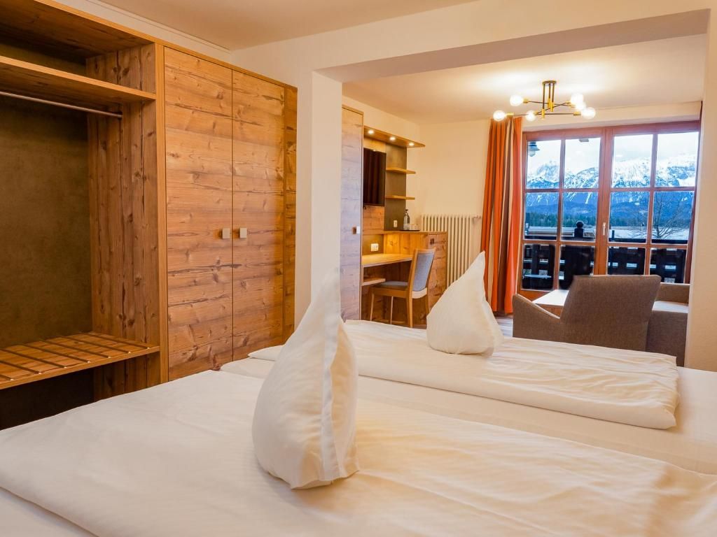 A hotel room with a wooden wardrobe, a desk with a chair, and a view of mountains through a large window.