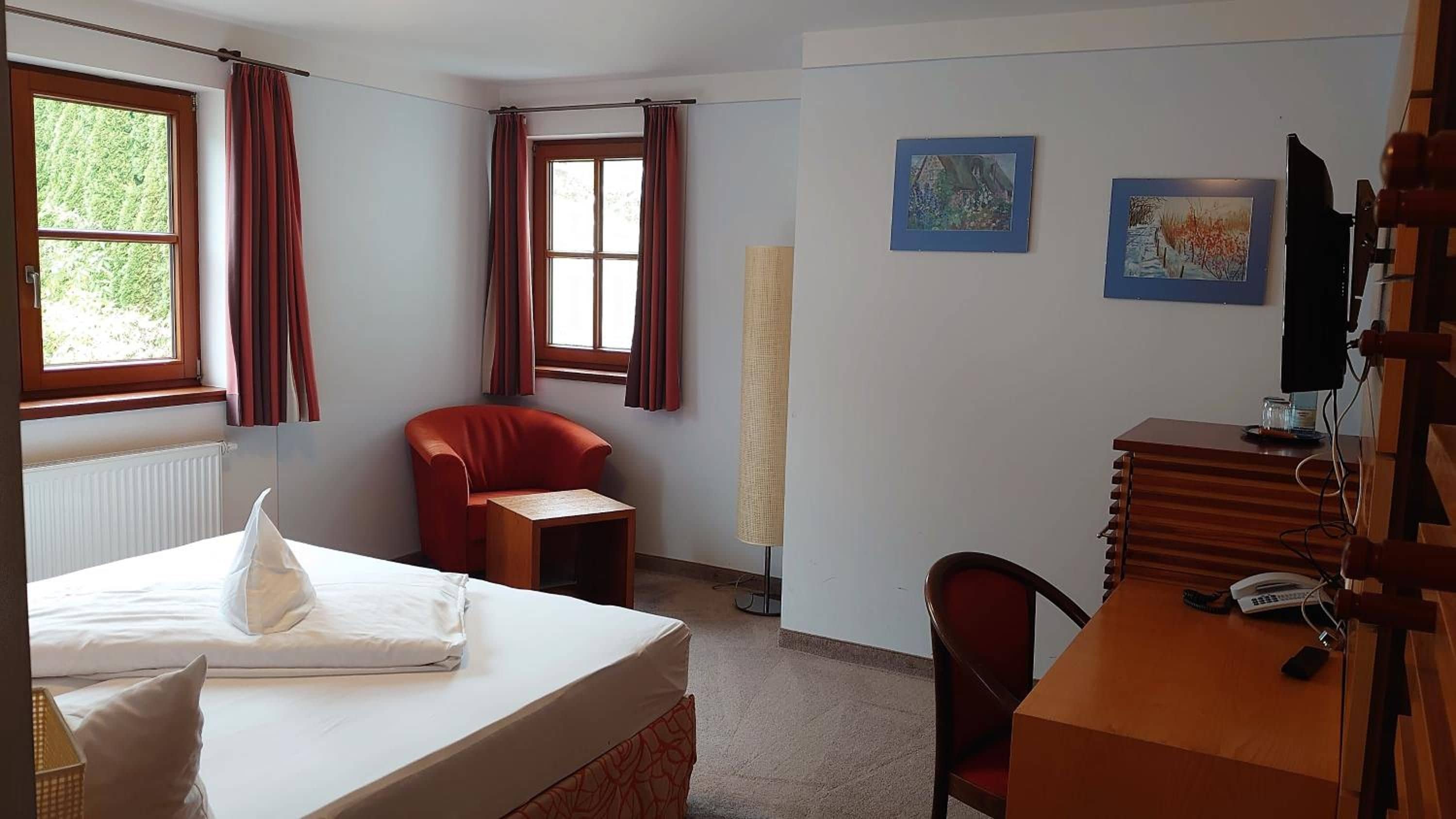 Ein Hotelzimmer mit einem Bett, einem Schreibtisch, einem Sessel und zwei mit Vorhängen versehenen Fenstern.