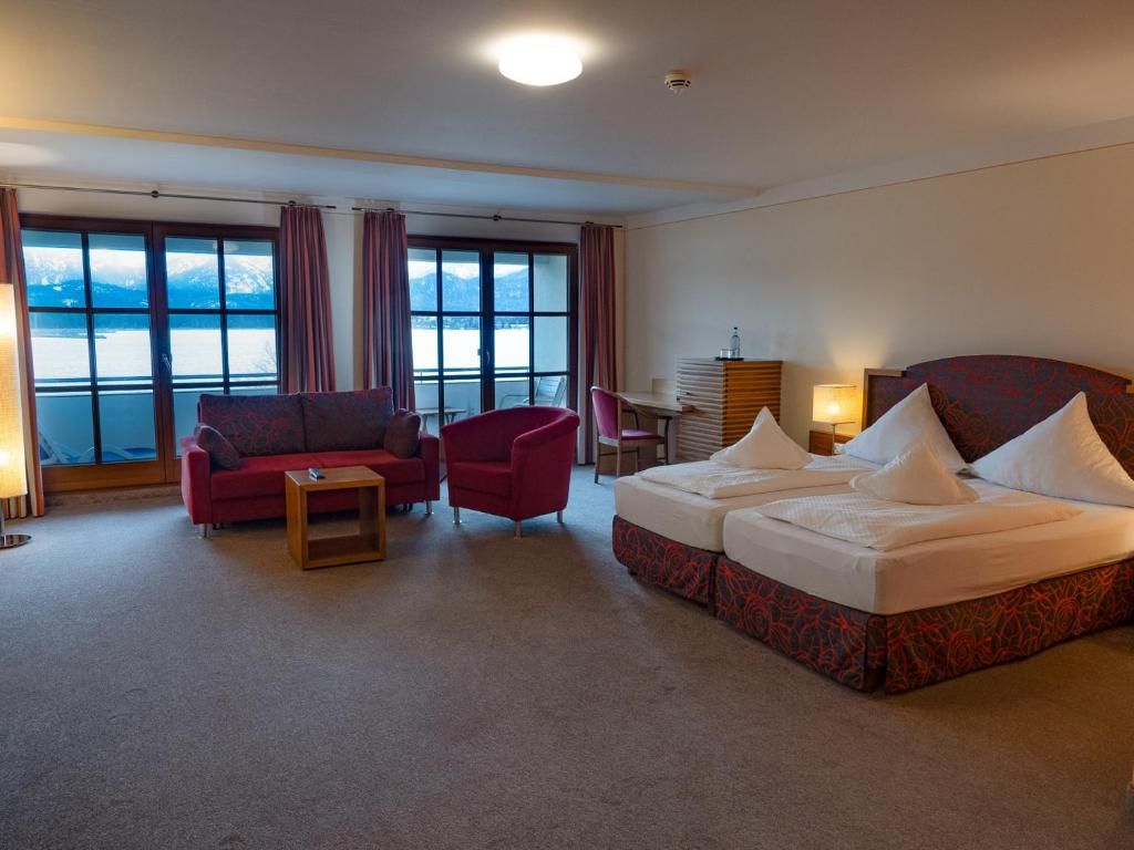 Ein Hotelzimmer mit Bett, einer Couch, einem Sessel, einem kleinen Tisch und Blick auf ein Gewässer durch große Fenster.
