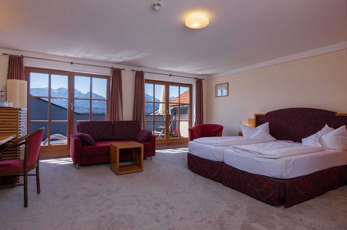 A hotel room with a bed, two red sofas, a small table, and a view of mountains through large windows.