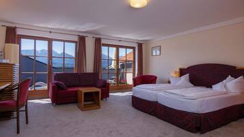 A hotel room with a bed, two red sofas, a small table, and a view of mountains through large windows.