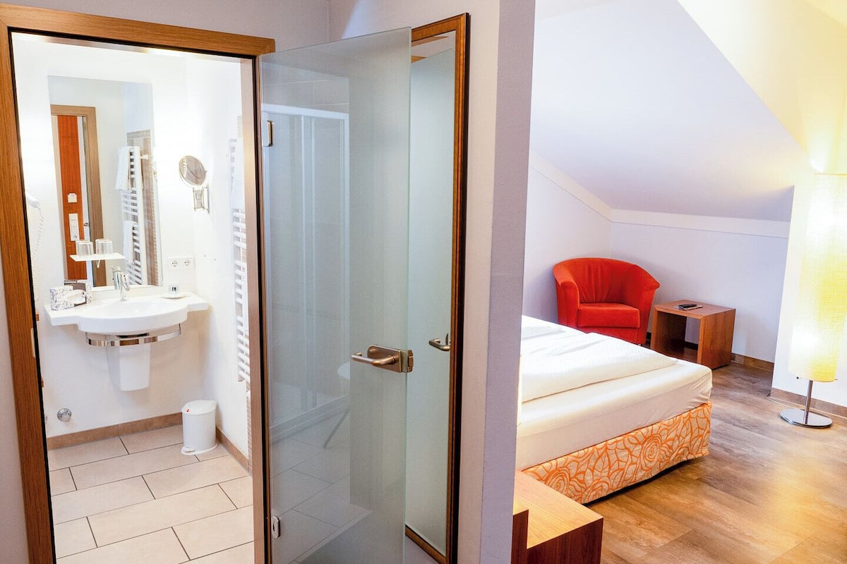 A modern hotel room with a glass shower door, a white sink, a red chair, and a bed with an orange patterned bedspread.