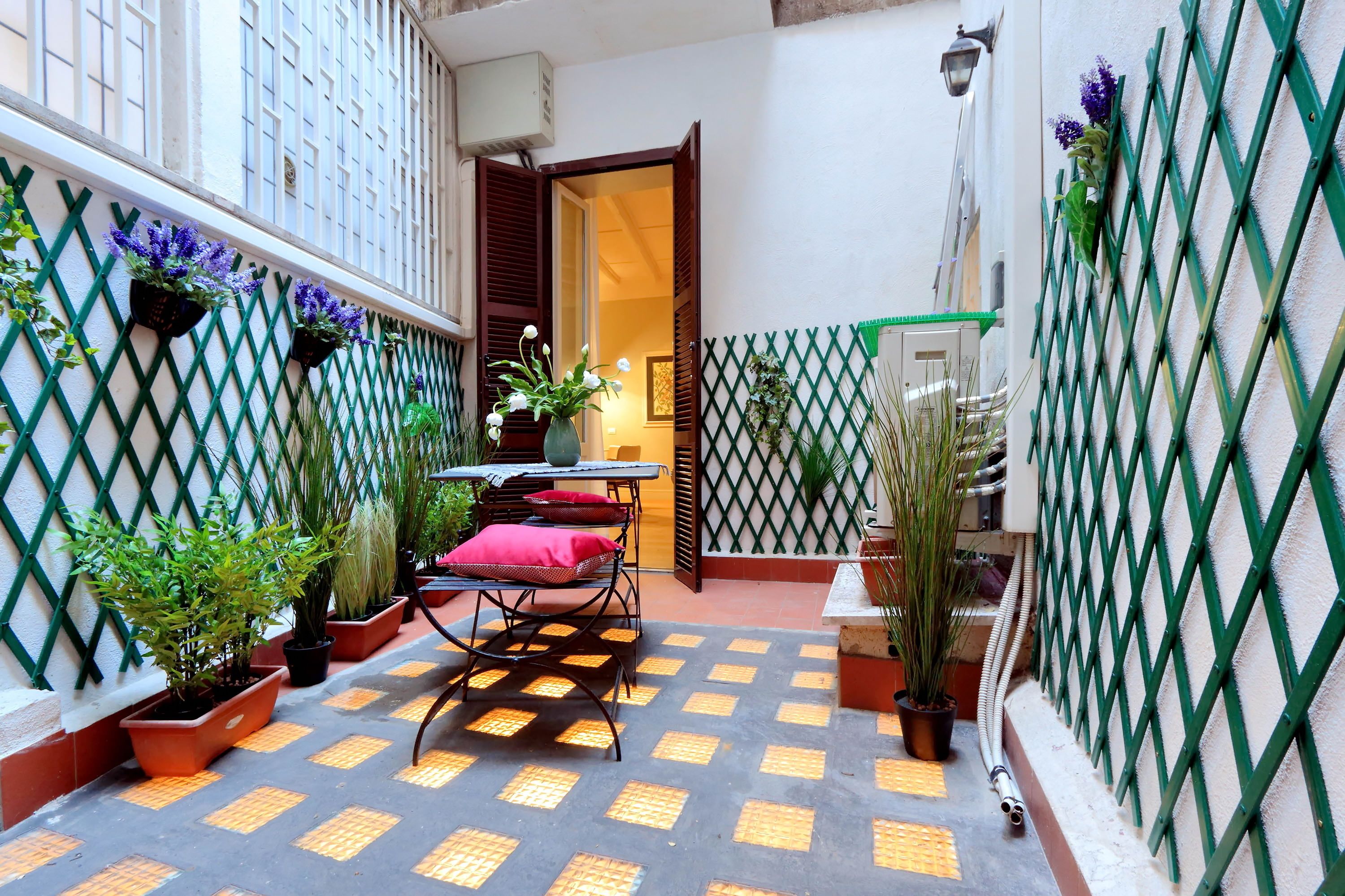 Une petite cour avec une table et des chaises, entourée de plantes en pot et d’une clôture en treillis.