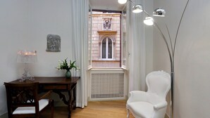 A room with a wooden desk, a white chair, a lamp, a vase with flowers, and a view of a building through a window with white curtains. - Farnese Charme - My Extra Home (Rome)