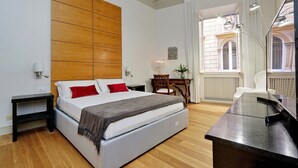 A modern bedroom with a wooden headboard, a bed with white linens and red pillows, a desk with a mirror, and a view of a building through a window. - Farnese Charme - My Extra Home (Rome)