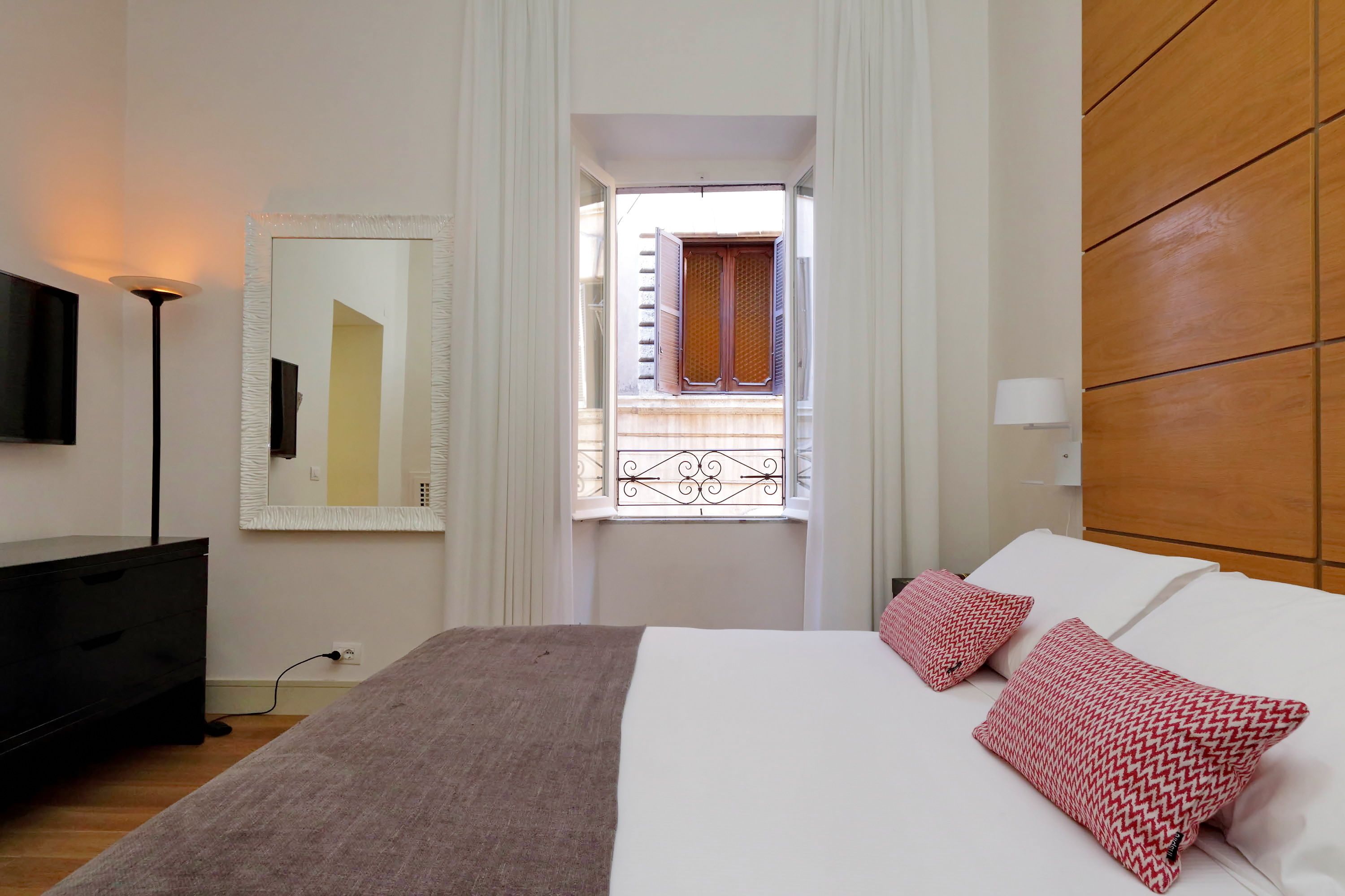 Ein Schlafzimmer mit Bett, Nachttischlampen, einem Schminktisch und einem Fenster mit Vorhängen, mit Blick nach draußen.