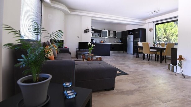Una sala de estar moderna con un sofá, una mesa de centro y una planta en maceta. Hay una zona de comedor con una mesa y sillas. La habitación tiene un ventanal grande y una cocina al fondo.