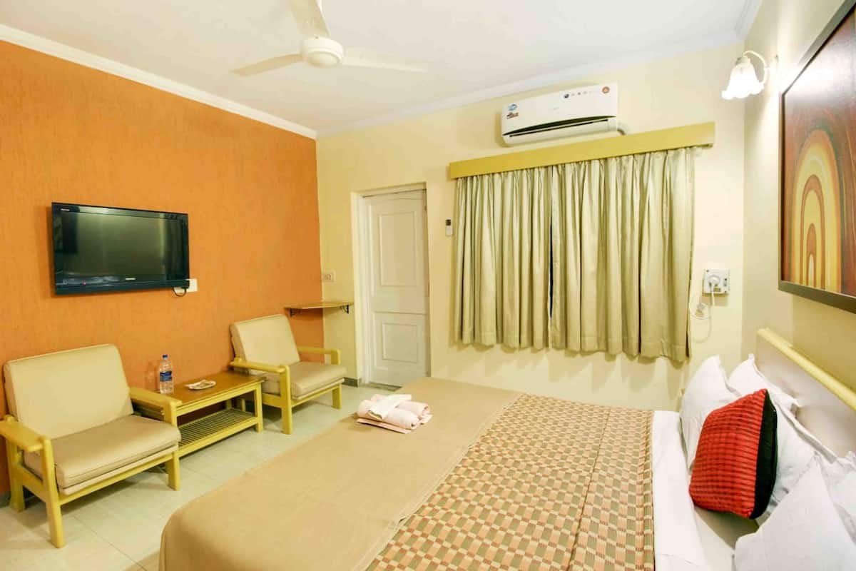 A hotel room with a bed, two chairs, a small table, a TV, an air conditioner, and a door.