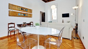 Un comedor con una mesa redonda, sillas transparentes, piso de madera, sofá blanco, televisor y techo con vigas a la vista.