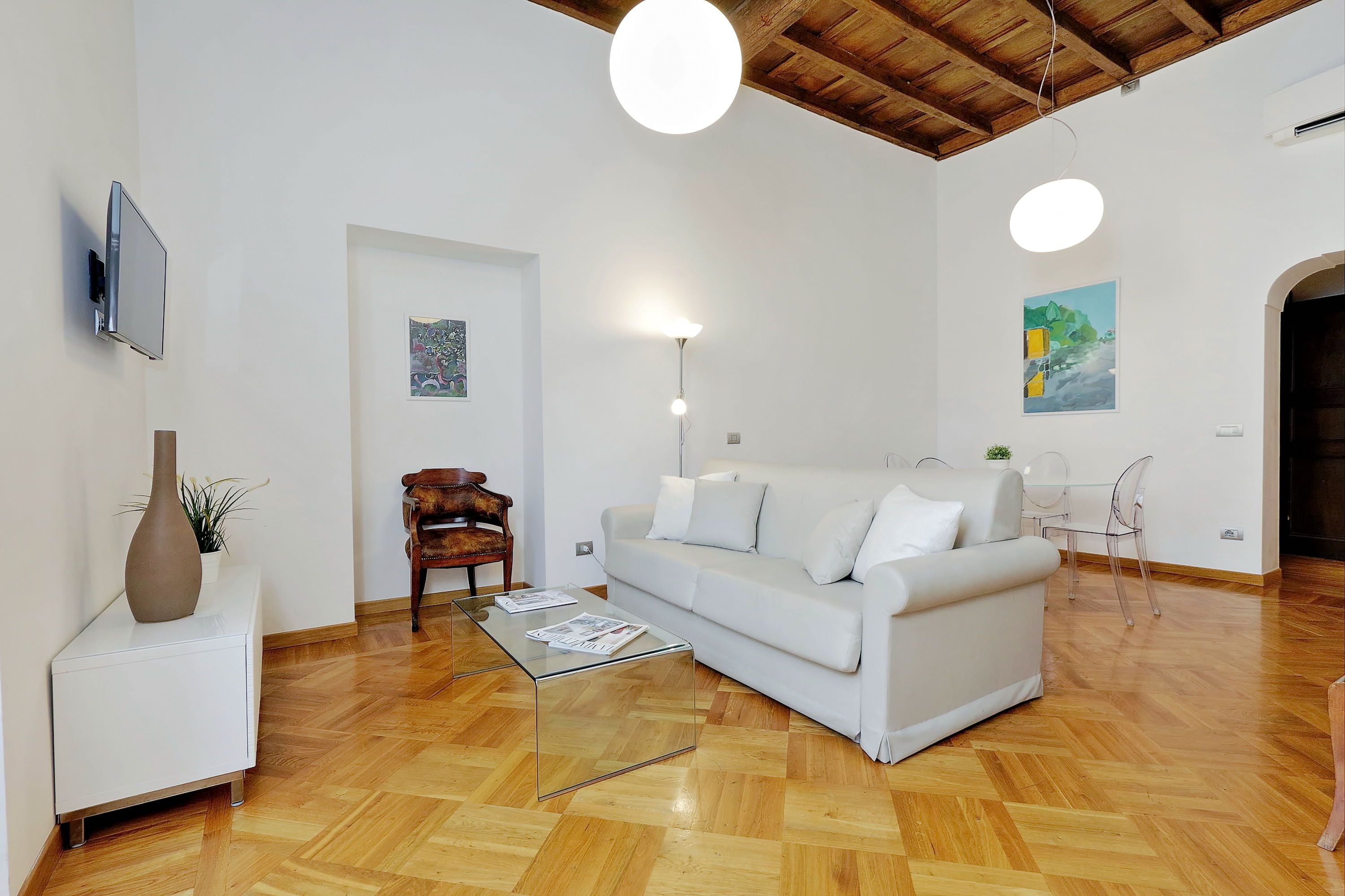 A modern living room with a white sofa, a glass coffee table, a wooden chair, and a TV mounted on the wall.
