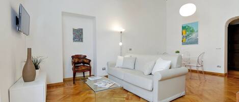 A modern living room with a white sofa, a glass coffee table, a wooden chair, and a TV mounted on the wall.