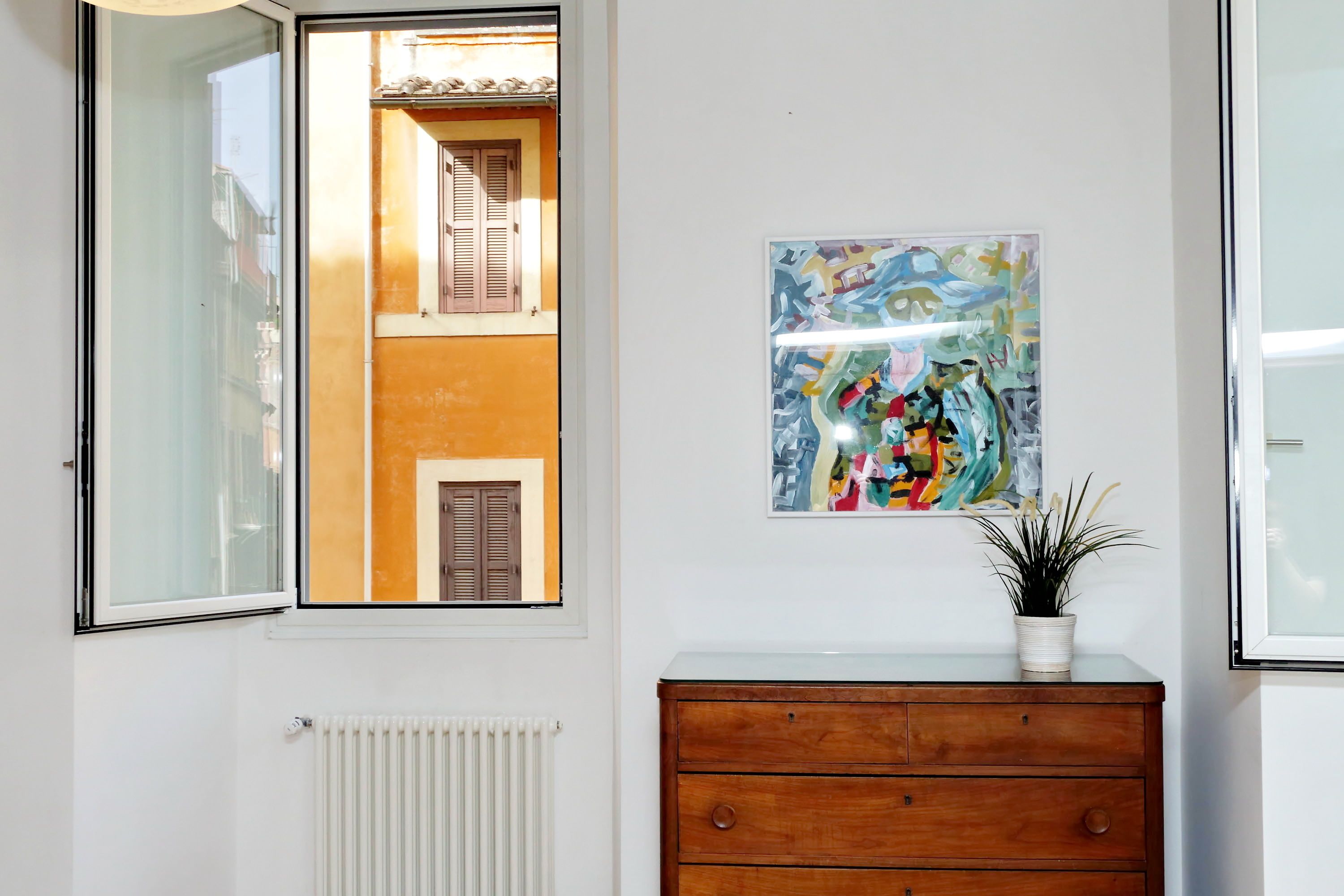 Une pièce avec une fenêtre, un radiateur, une commode en bois et un tableau abstrait encadré.