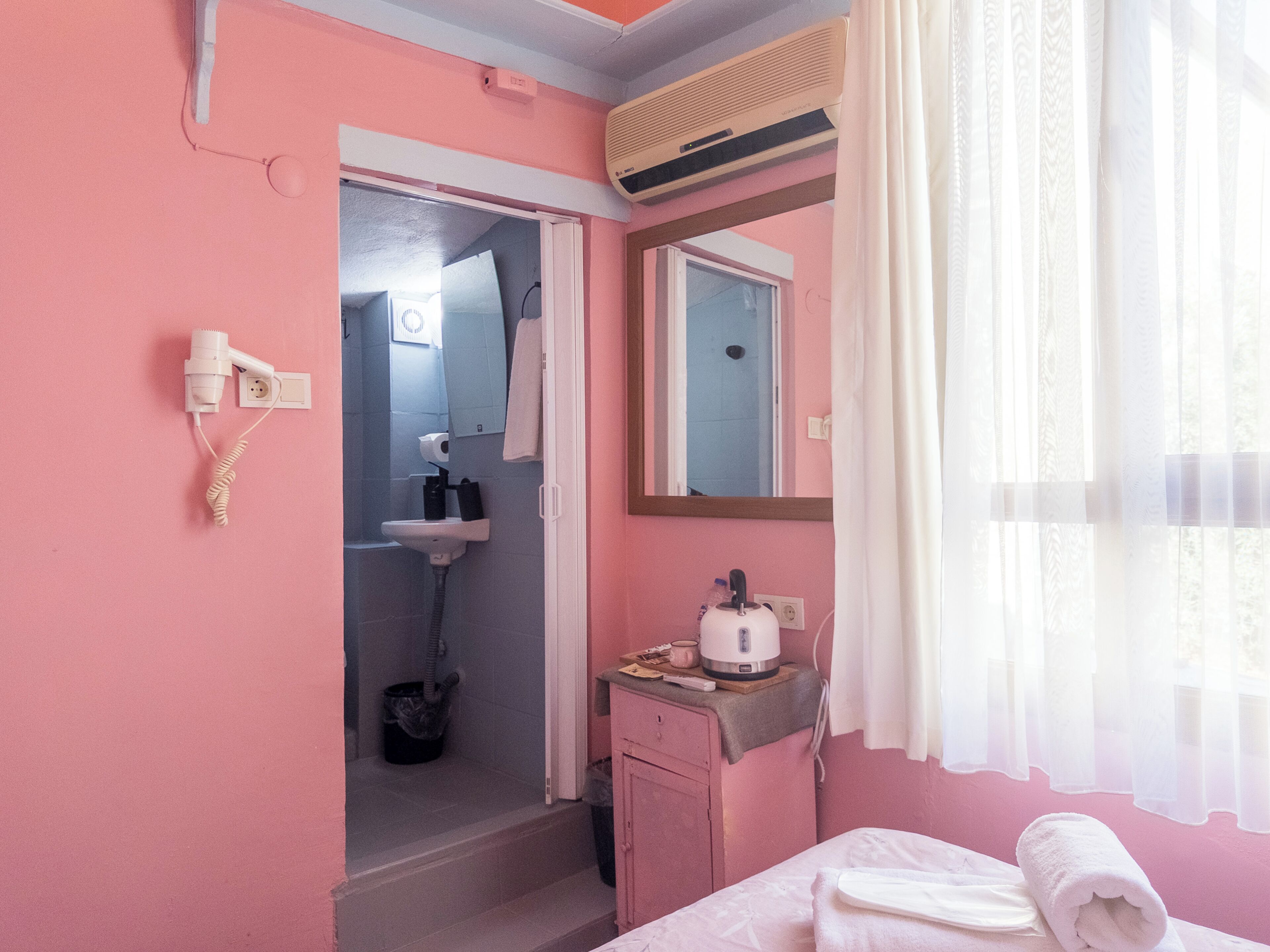 Una habitación rosa con una cama, una mesita de noche con una tetera, un espejo y una ventana con cortinas translúcidas.