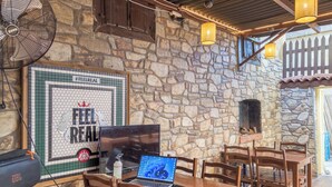 A stone-walled outdoor seating area with a laptop on a table, wooden chairs, a fan, and string lights. - Tarihi Rum Konağı Antikhan (Ayvalik)