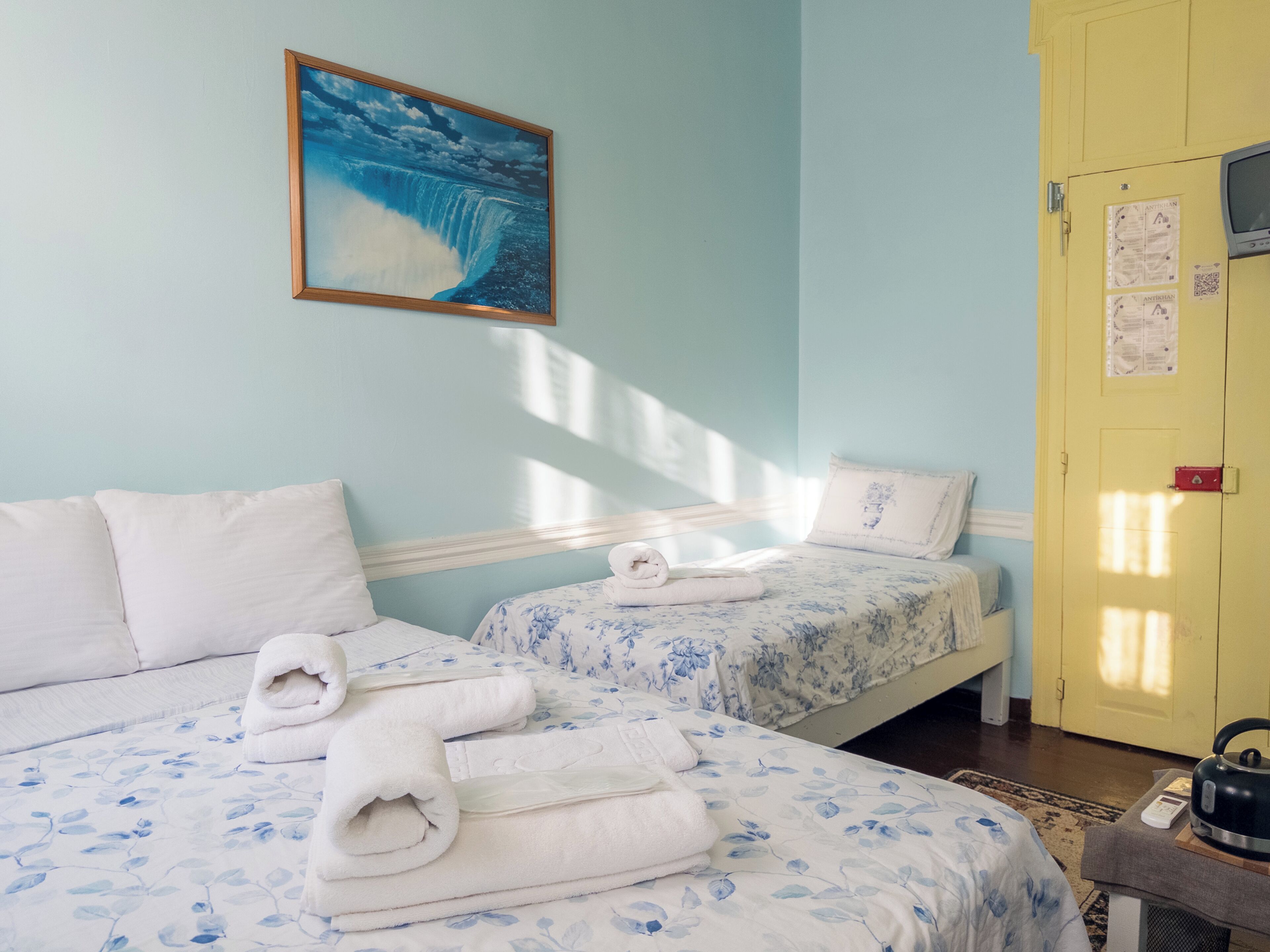 Una habitación con dos camas, un cuadro de una cascada, una puerta amarilla y un hervidor sobre una mesa.