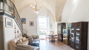 A spacious living room with a high arched ceiling, a staircase with black railings, a chandelier, a dining area with a round table, and a large wooden cabinet with glass doors. - B&B Donna Lina (Matera)