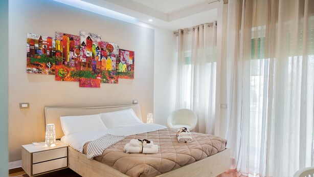 Un dormitorio moderno con una cama grande, una mesita de noche, una silla y una ventana con cortinas traslúcidas.