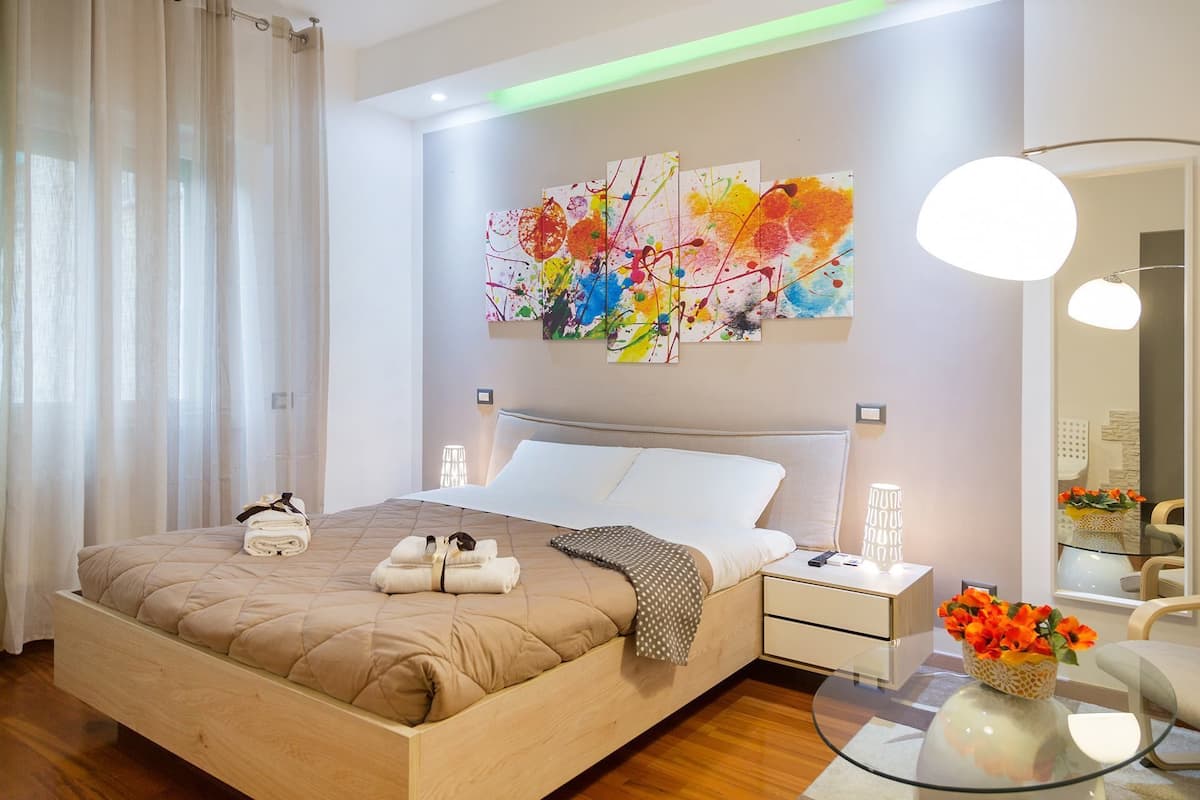 A modern hotel room with a large bed, a bedside table, a glass coffee table, and a vibrant abstract painting on the wall.