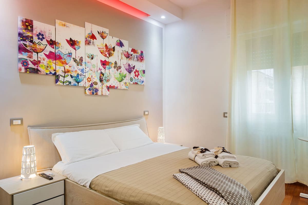 A neatly made bed with a beige bedspread, a pillow, and a blanket. There are two bedside tables with lamps and a remote control on one. A set of towels is folded on the bed.