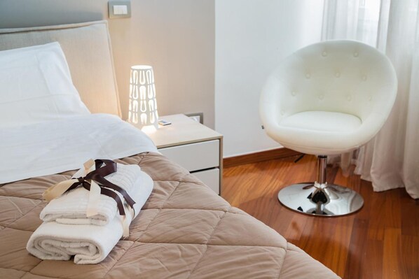 Una cama bien hecha con una toalla doblada y un almohadón, una silla blanca de diseño moderno y una mesita de noche con una lámpara.