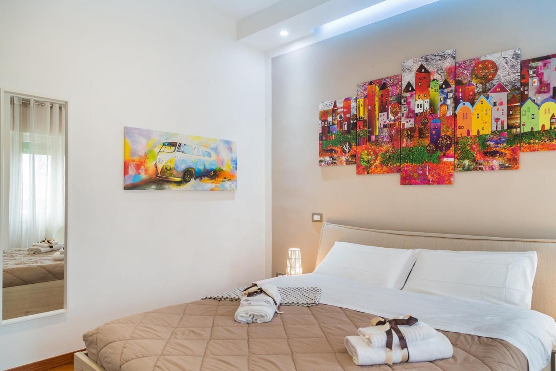 Quarto de hotel com uma cama, um quadro de um carro e um mural colorido de casas.