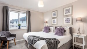 A bedroom with a large bed, a chair, a bedside table, a lamp, and framed pictures on the wall. - Bay View - Donnini Apartments (Ayr)