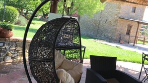 A hanging chair with a cushion, wicker furniture, and a stone building in the background. - Azienda Agrituristica I Tre Casali (Magione)