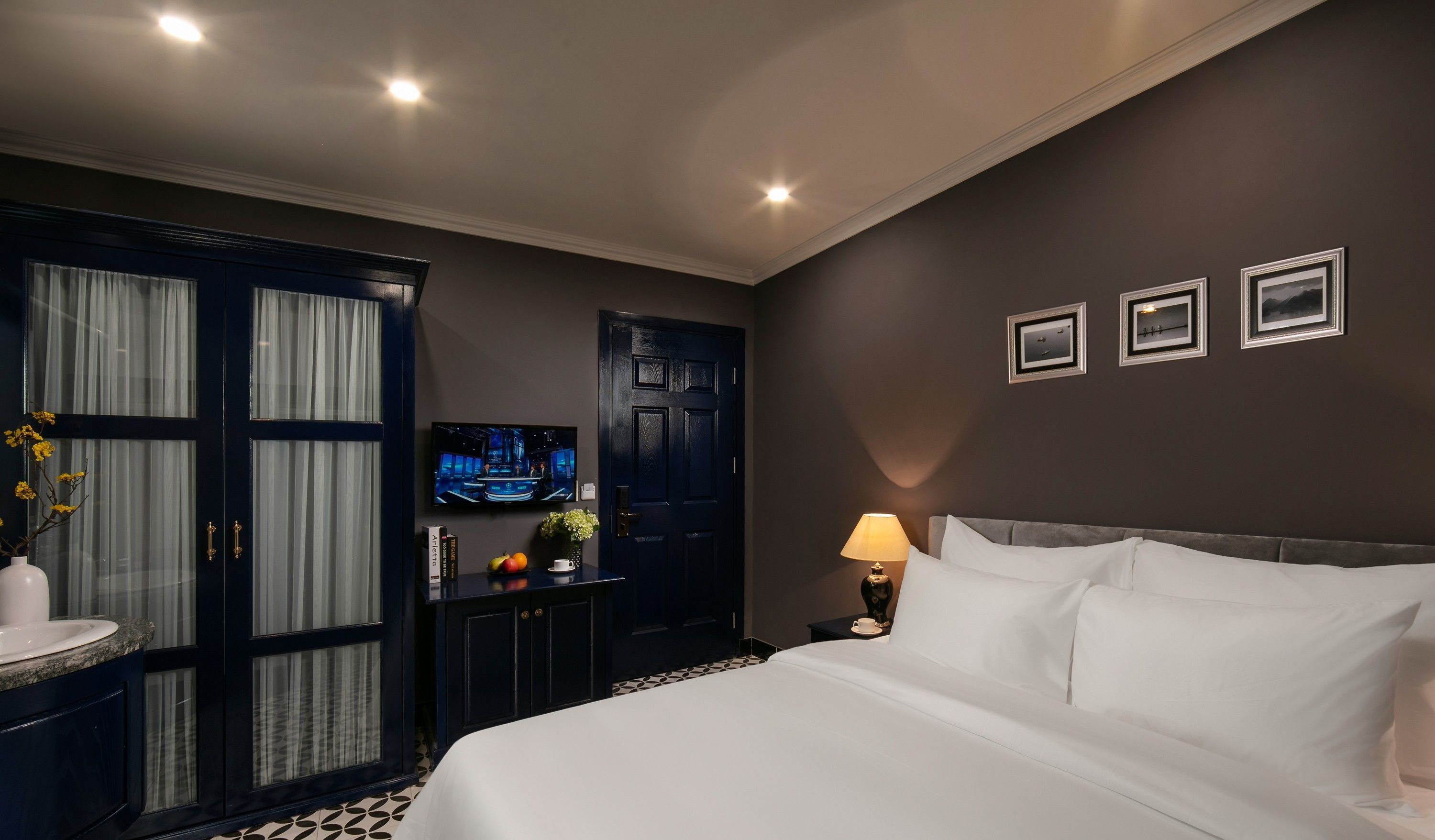 A hotel room with a large bed, a TV mounted on the wall, a desk with a lamp, and a wardrobe.