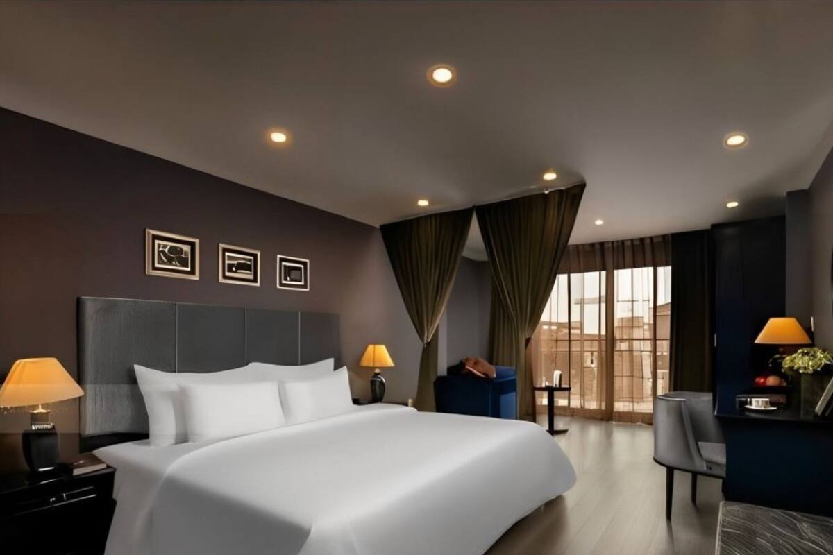 A modern hotel room with a large bed, two bedside tables with lamps, a desk with a chair, a large window with curtains, and a wall-mounted TV.