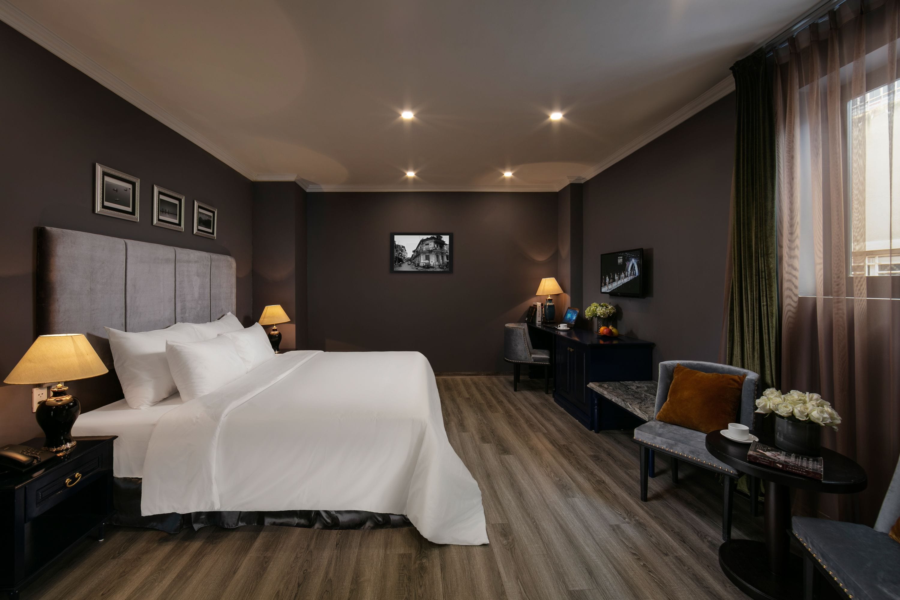 Ein Hotelzimmer mit einem großen Bett, Nachttischlampen, einem Schreibtisch mit Stuhl, einem kleinen Tisch mit Blumenvase und einem Bild an der Wand.