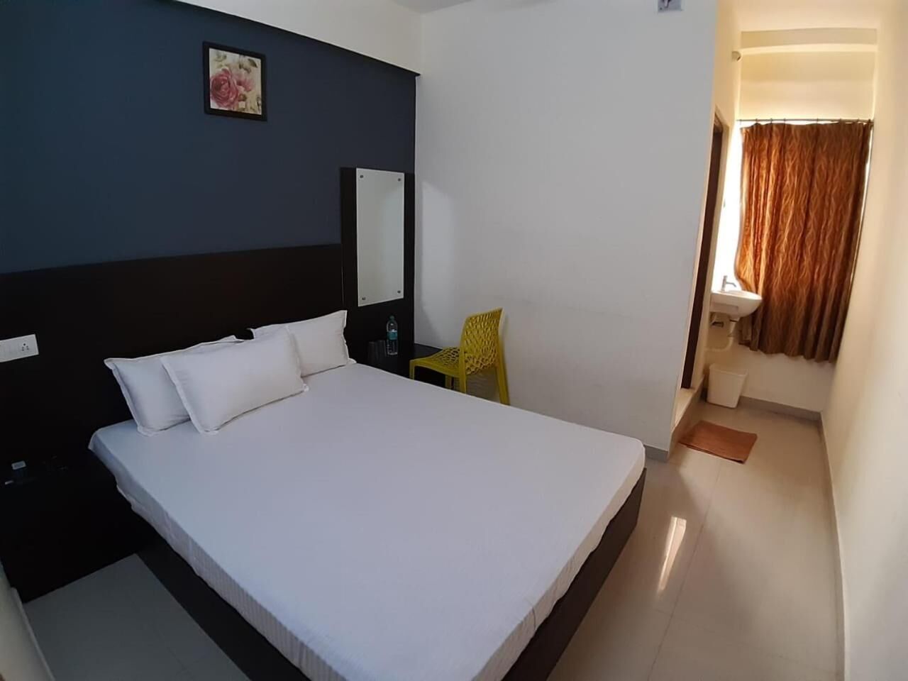 A neatly made bed in a room with a dark wall, a small yellow chair, and a bathroom visible through an open door.