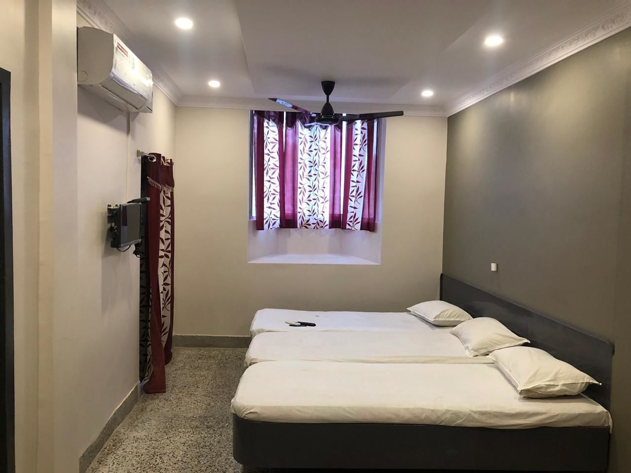 Habitación de hotel con dos camas, ventilador de techo, unidad de aire acondicionado y ventana con cortinas estampadas.