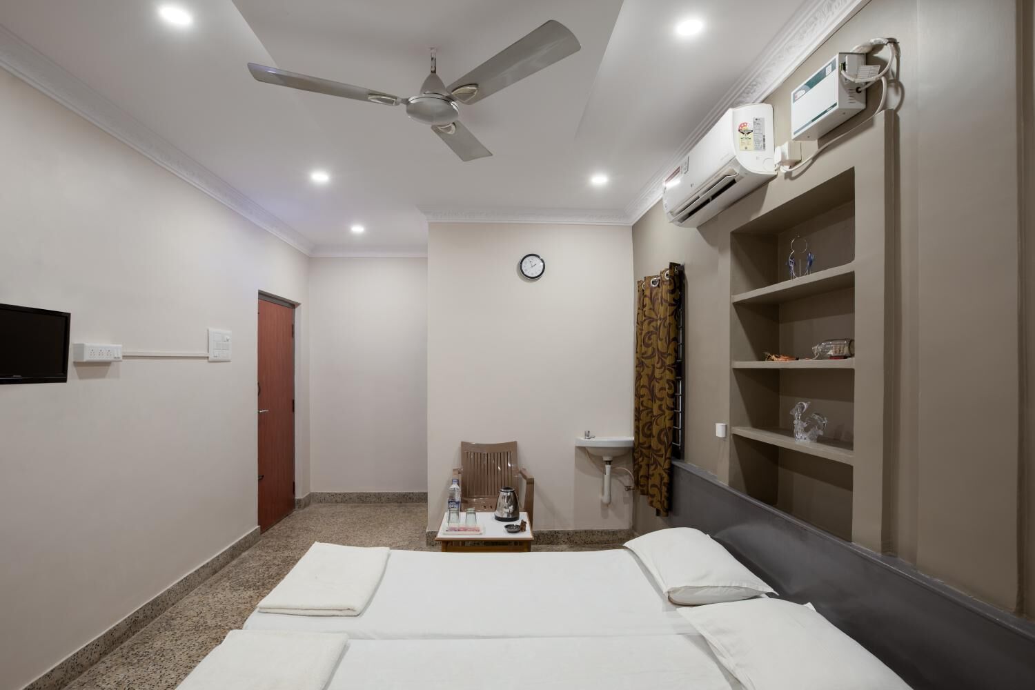 Ein Hotelzimmer mit Bett, Schreibtisch, Stuhl, Waschbecken, einer Uhr und einem Deckenventilator.