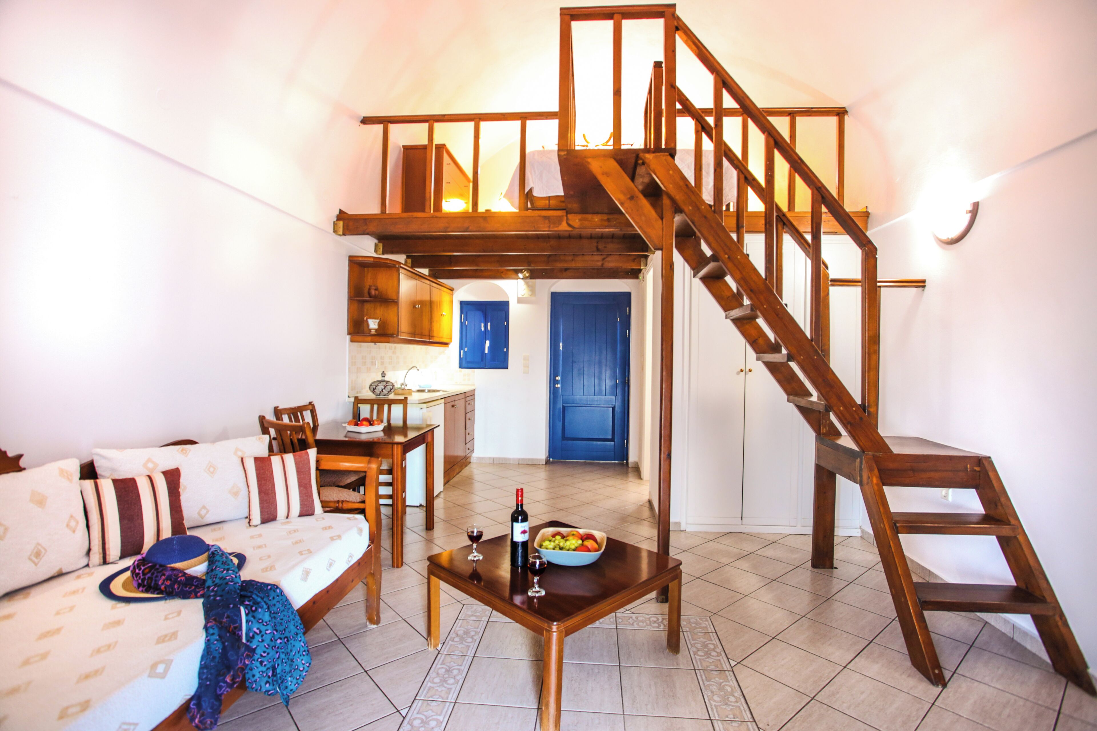 A spacious living area with a wooden staircase leading to an upper level. A dining table with chairs and a kitchen area are visible in the background.