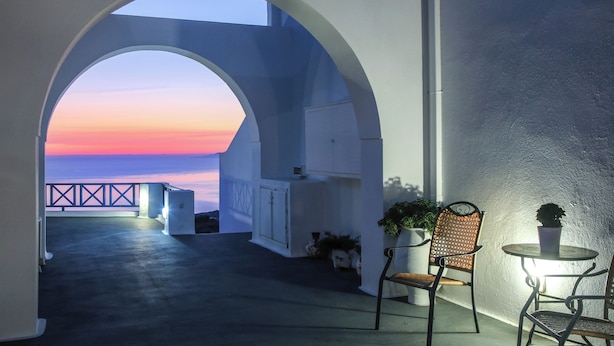An outdoor terrace with a view of the sunset, a white building, and two chairs with potted plants.