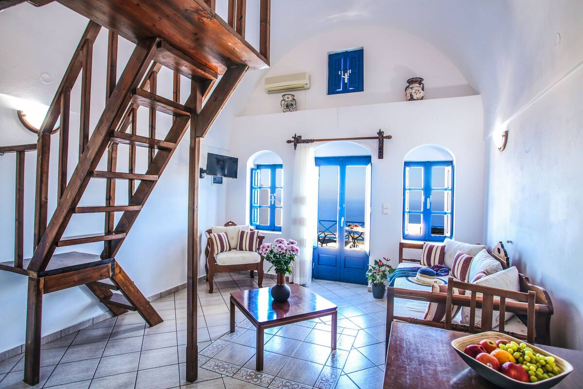 A spacious living room with a wooden staircase, a sofa set, a dining table with fruit, and a view through blue doors.