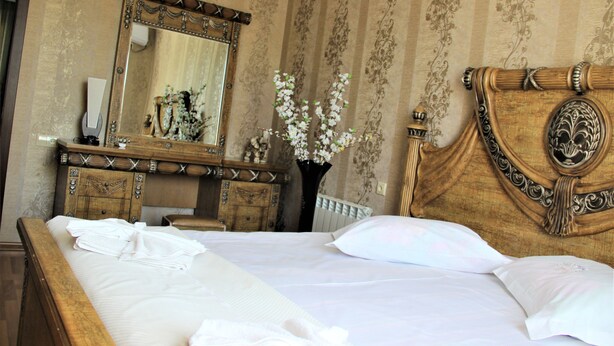 A neatly made bed with white linens, a wooden headboard with intricate carvings, a large mirror above the dresser, and a vase with white flowers on the dresser. - Hotel Oniks (Kutaisi)