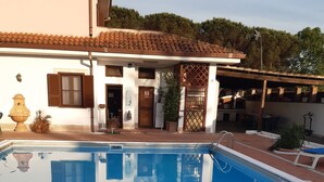 Une piscine à l’eau d’un bleu cristallin, entourée d’un bâtiment blanc doté d’un toit en tuiles et d’une pergola en bois.