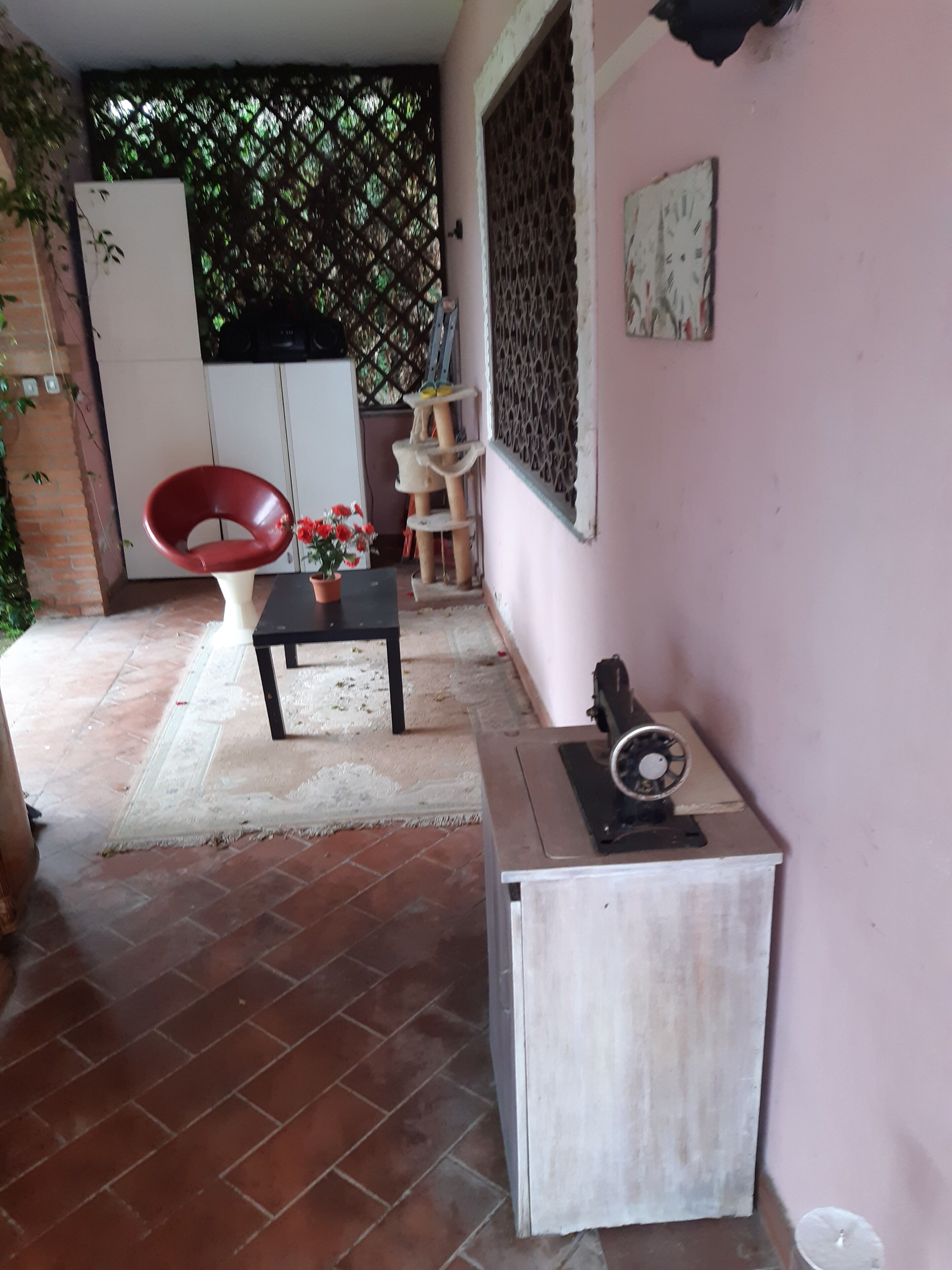 Una habitación pequeña con piso de baldosas, una silla roja, una mesa con un florero y una máquina de coser, además de un armario blanco.