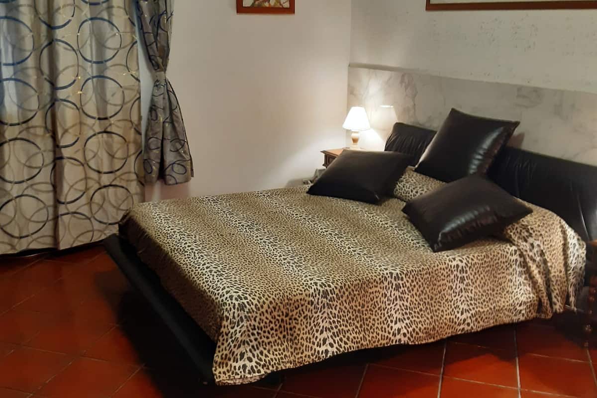 A bedroom with a bed, a nightstand, a lamp, a painting, and a window with curtains.