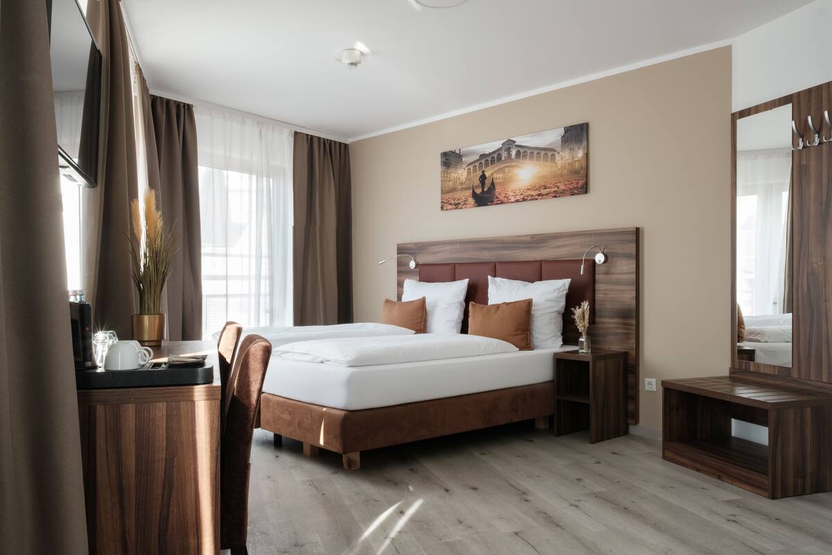 A hotel room with a large bed, a wooden desk, a chair, a bedside table, and a mirror.