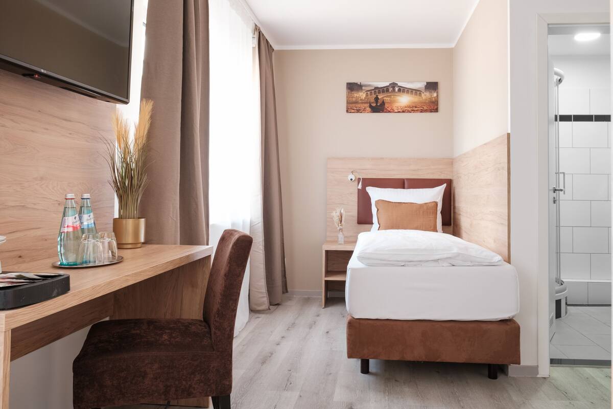A hotel room with a bed, a desk, a chair, a TV, a picture on the wall, and a shower area.