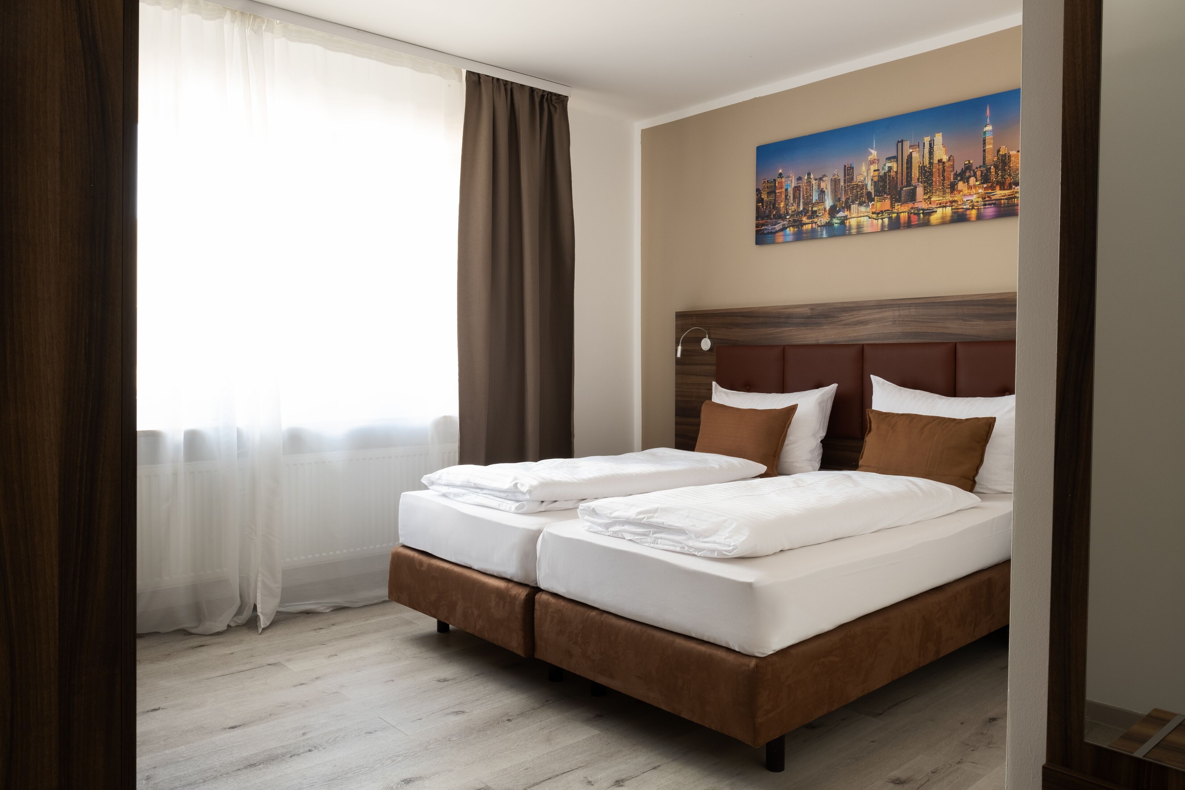 Een tweepersoonsbed met een houten hoofdbord, wit beddengoed en bruine kussens in een kamer met licht houten vloeren en een stadsgezichtschilderij aan de muur.