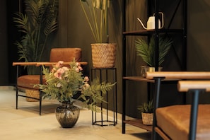 A modern living room with a brown leather sofa, a glass vase with flowers, a woven basket, and a small potted plant on a black metal stand. - The sQuare Hotel  (Frankfurt)