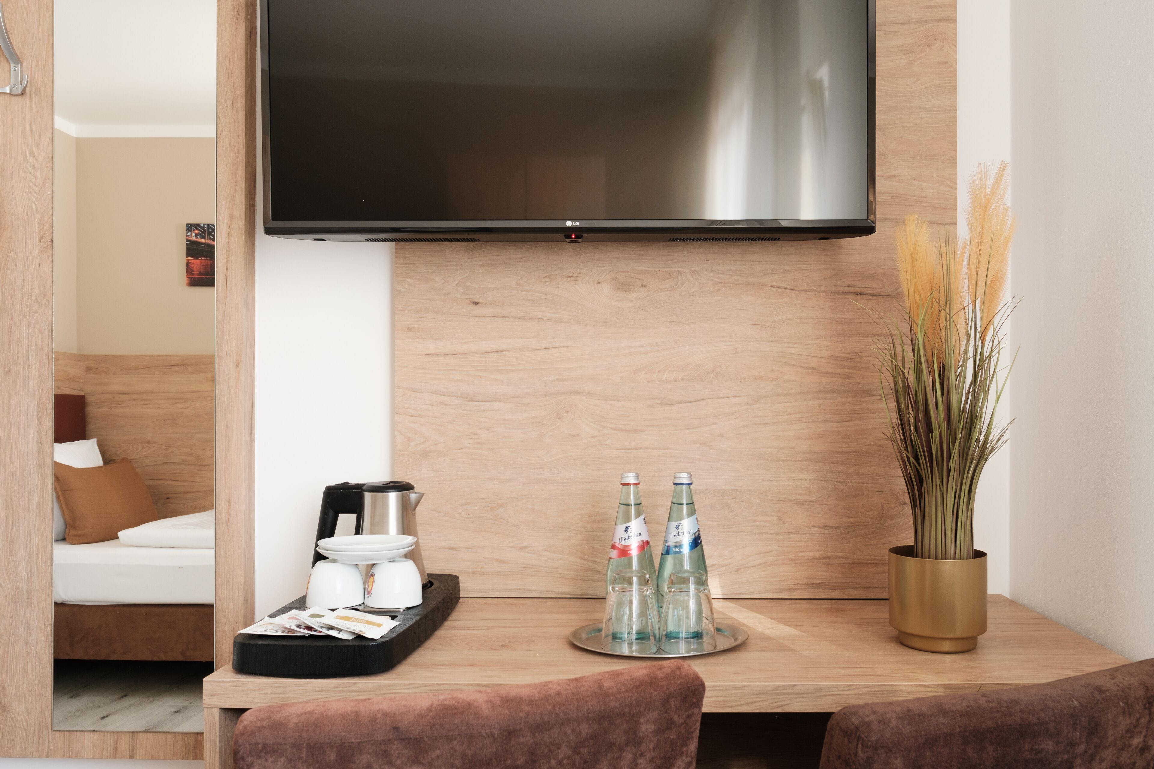 A modern hotel room with a flat-screen TV mounted on the wall, a wooden coffee table with a coffee maker, two bottles of water, and a potted plant.
